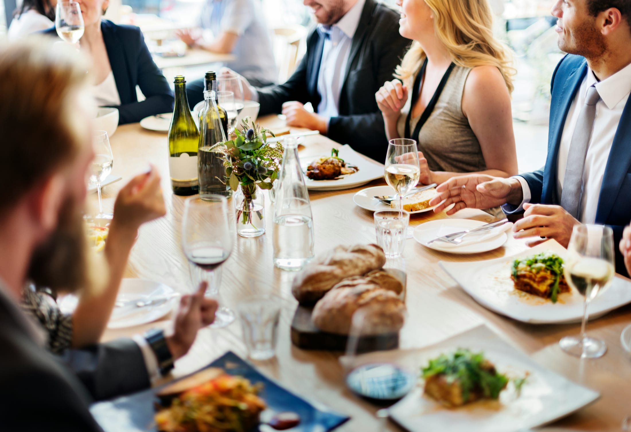 Guests in business jackets talking around a daylit restaurant table set with white wine, bread, flowers, and plated lunch